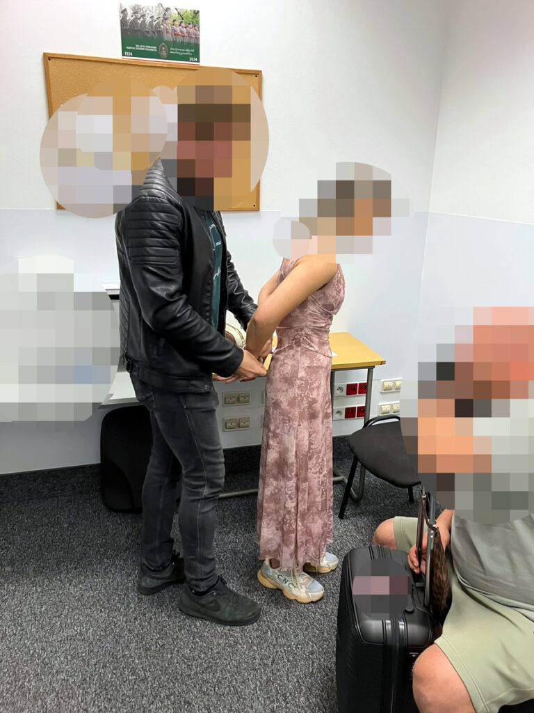 Mężczyzna w czarnej skórzanej kurtce zakłada kajdanki na ręce kobiety w różowej sukience w pomieszczeniu biurowym.
