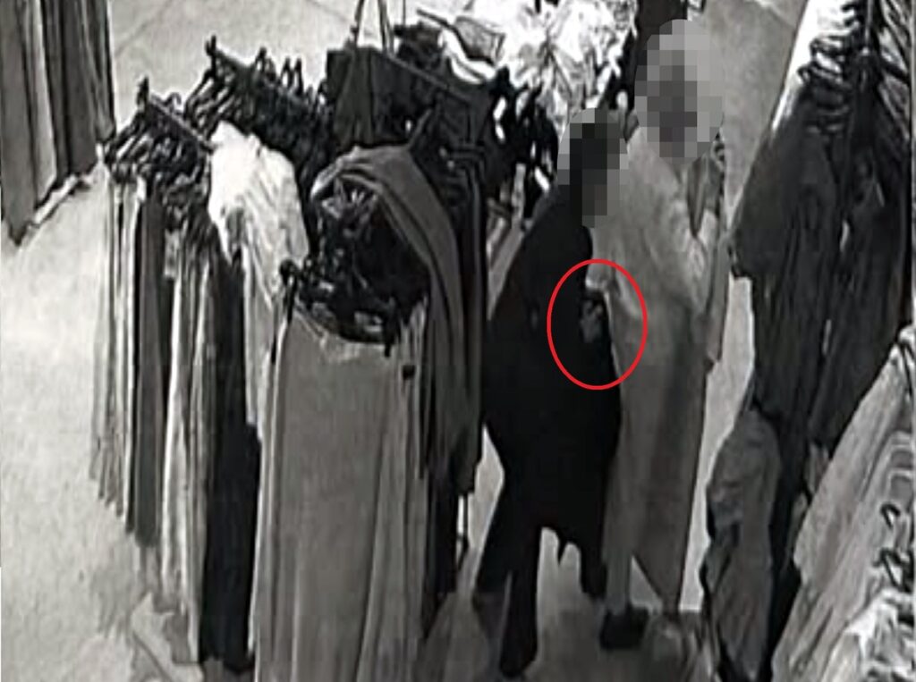Osoba w czarnym płaszczu stoi obok innej osoby, trzymając coś w ręku, w sklepie odzieżowym.