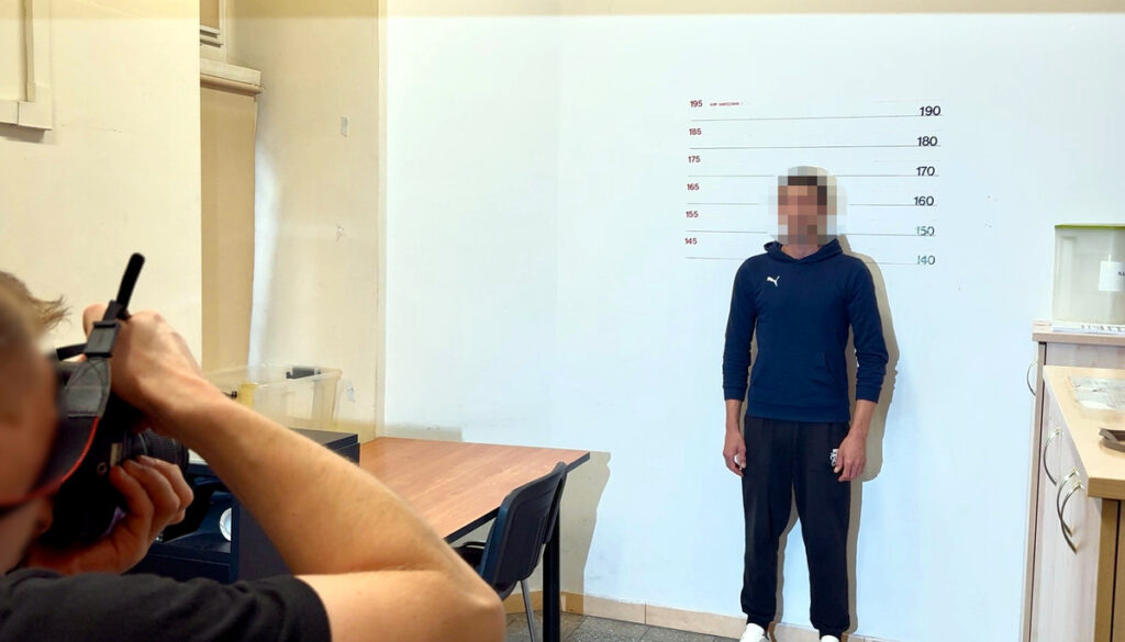 Mężczyzna w dresie stoi przy ścianie z oznaczeniami wysokości, podczas gdy fotograf robi mu zdjęcie.