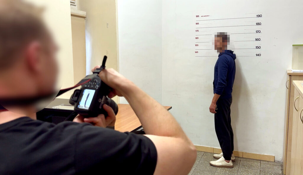Mężczyzna w niebieskiej bluzie stoi przy ścianie z podziałką wysokości, podczas gdy fotografuje go inny mężczyzna.