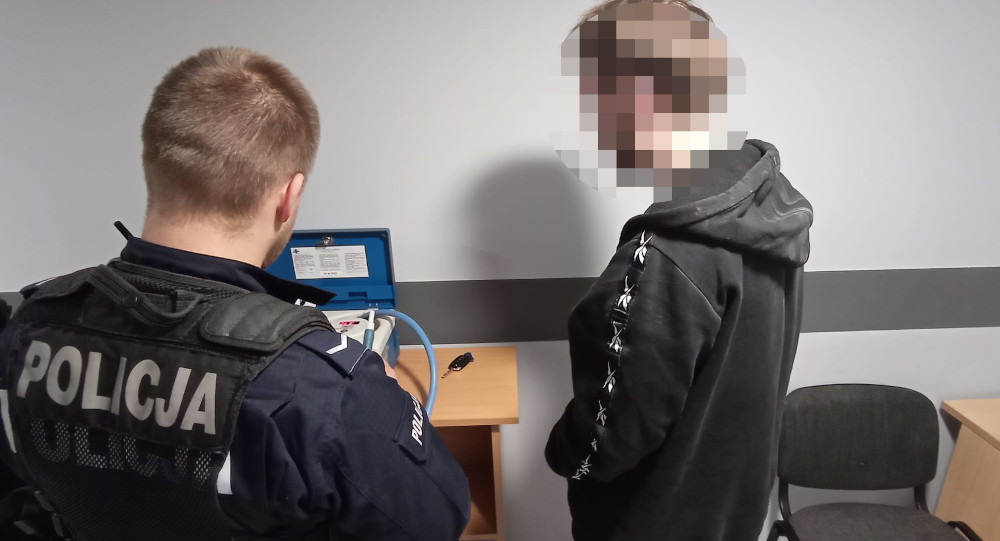 Policjant w mundurze wykonuje badanie alkomatem na miejscu, obok stoi mężczyzna w czarnej bluzie.