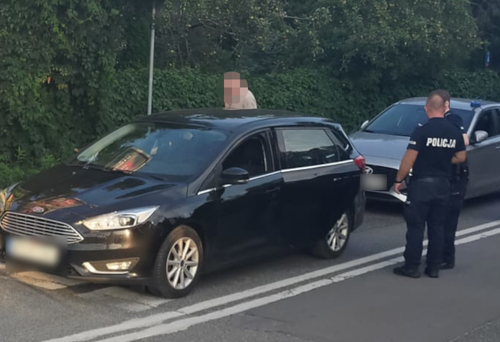 Mężczyzna stojący obok zatrzymanego czarnego samochodu, a dwóch policjantów rozmawia z kierowcą na poboczu drogi.