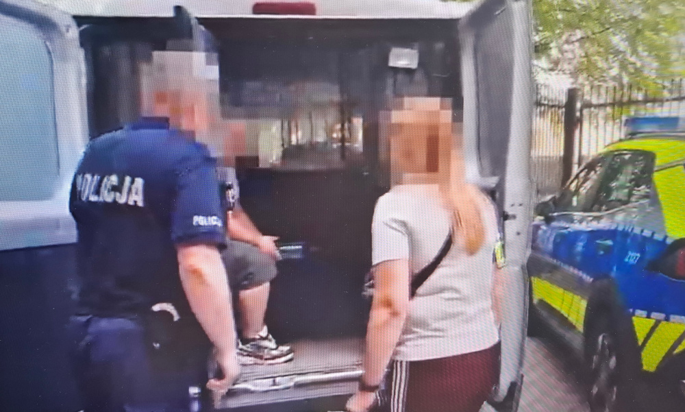 Mężczyzna w kajdankach wsiada do radiowozu, obok stoi policjant i kobieta. W tle widoczny policyjny samochód.