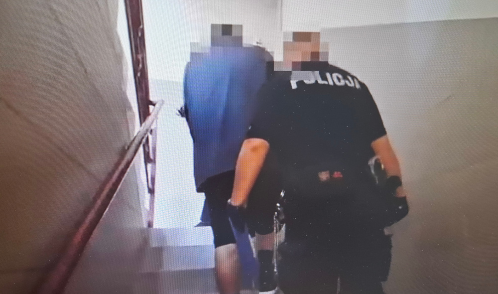 Mężczyzna w niebieskiej bluzie prowadzony przez policjanta po schodach w budynku mieszkalnym.