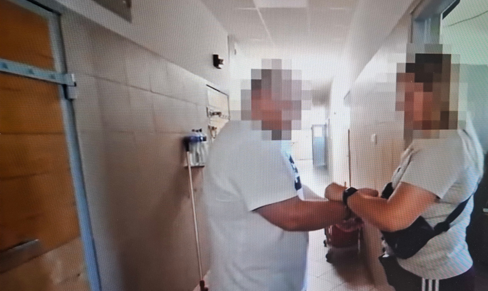 Mężczyzna w białej koszulce stoi w korytarzu, rozmawiając z policjantką, która trzyma go za rękę.