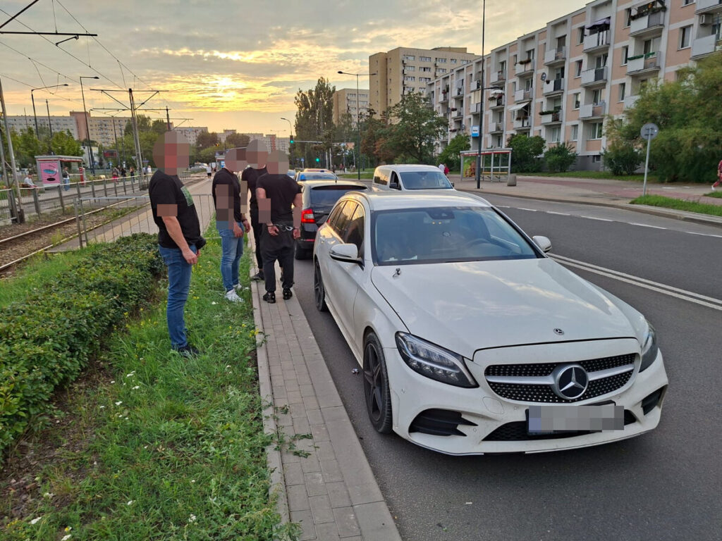 Grupa mężczyzn w czarnych koszulkach stoi obok białego samochodu marki Mercedes na ulicy, w tle widać budynki mieszkalne i tramwaj.