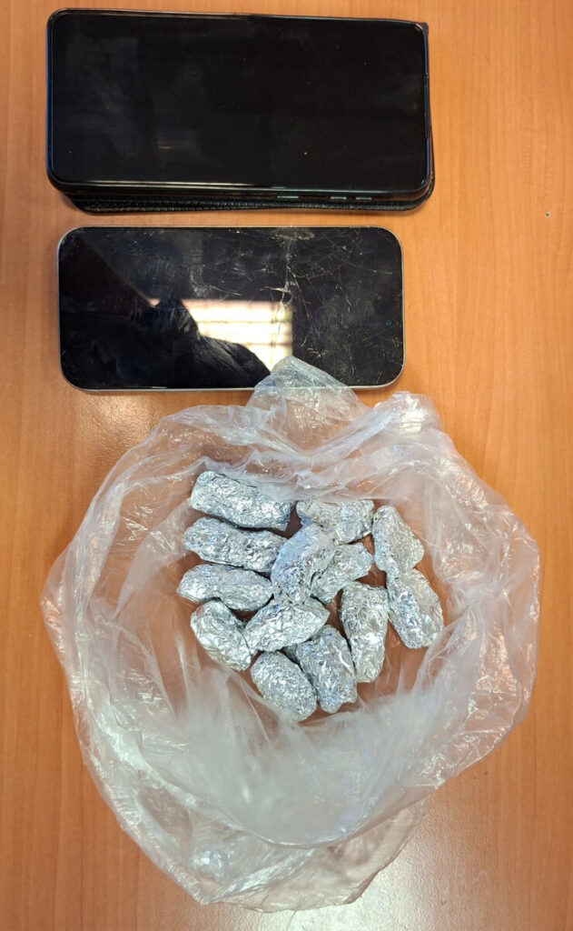 Dwa telefony komórkowe leżące na drewnianym stole obok torebki z zawiniętymi w folię aluminiową przedmiotami.