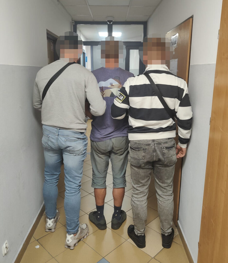 Trzech mężczyzn idzie korytarzem, jeden z nich jest prowadzony przez pozostałych. W tle widać drzwi i oświetlenie.