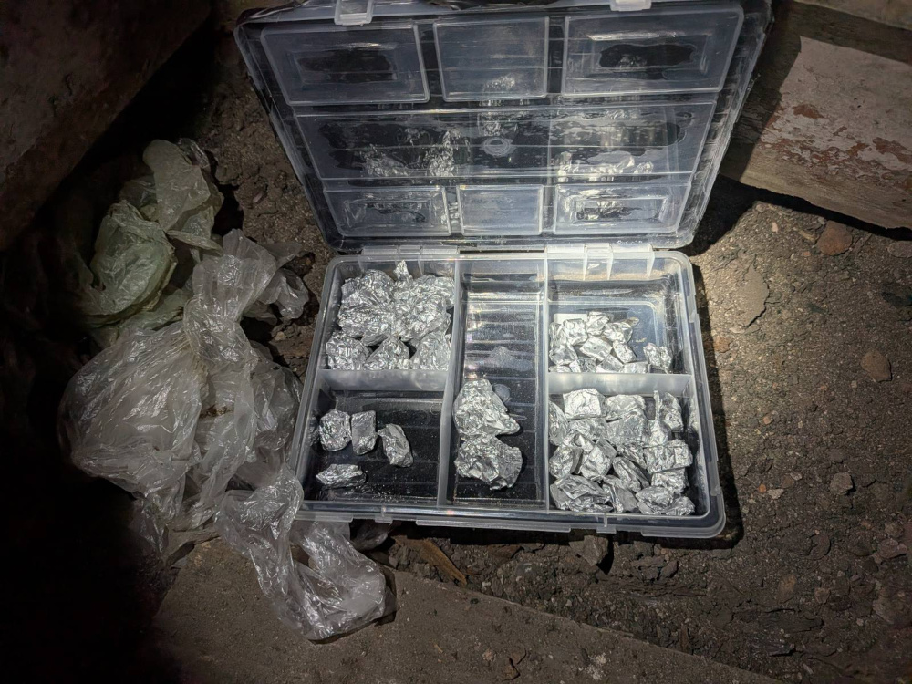 Otwarta plastikowa skrzynka z foliowymi pakunkami, wypełniona srebrnymi torebkami z substancjami, leży na ziemi.