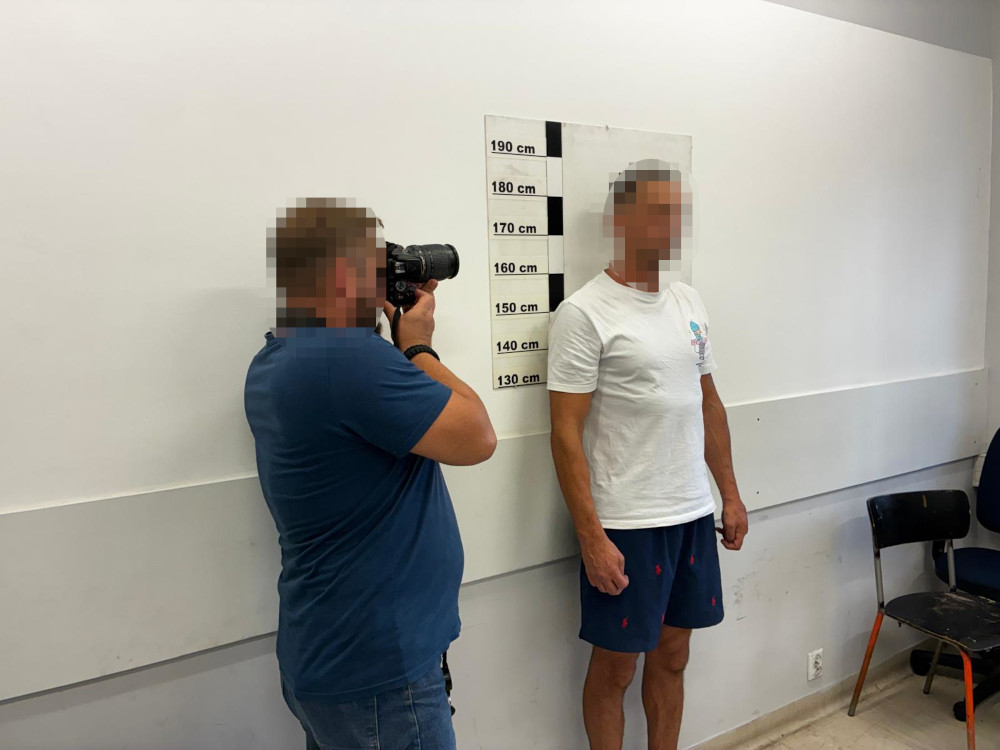 Mężczyzna w białej koszulce stoi przy ścianie z oznaczeniami wysokości, podczas gdy fotografuje go inny mężczyzna.