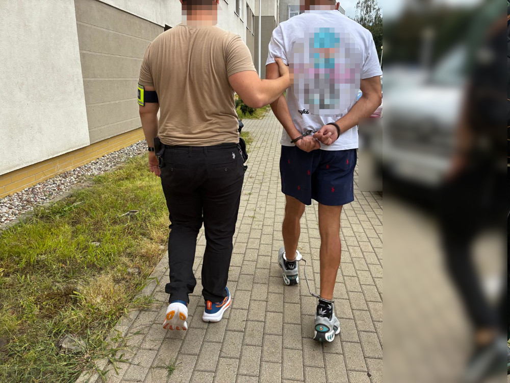 Funkcjonariusz policji prowadzi mężczyznę w kajdankach po chodniku w pobliżu budynku.