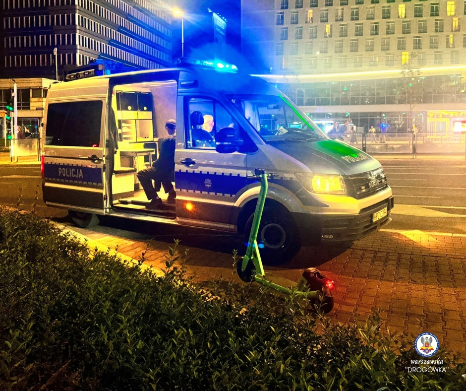 Policjant w mundurze siedzi w otwartym radiowozie, obok leży zielona hulajnoga na oświetlonym chodniku w nocy.
