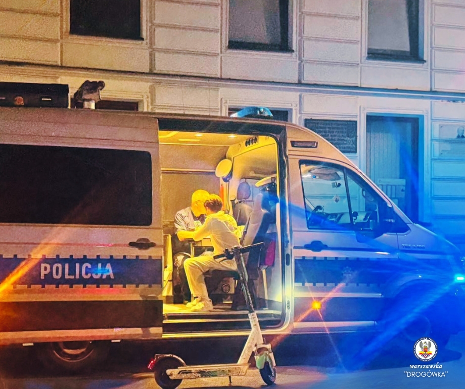 Policjant i mężczyzna siedzący w radiowozie, oświetlonym niebieskimi i żółtymi światłami, na tle miejskiego budynku.