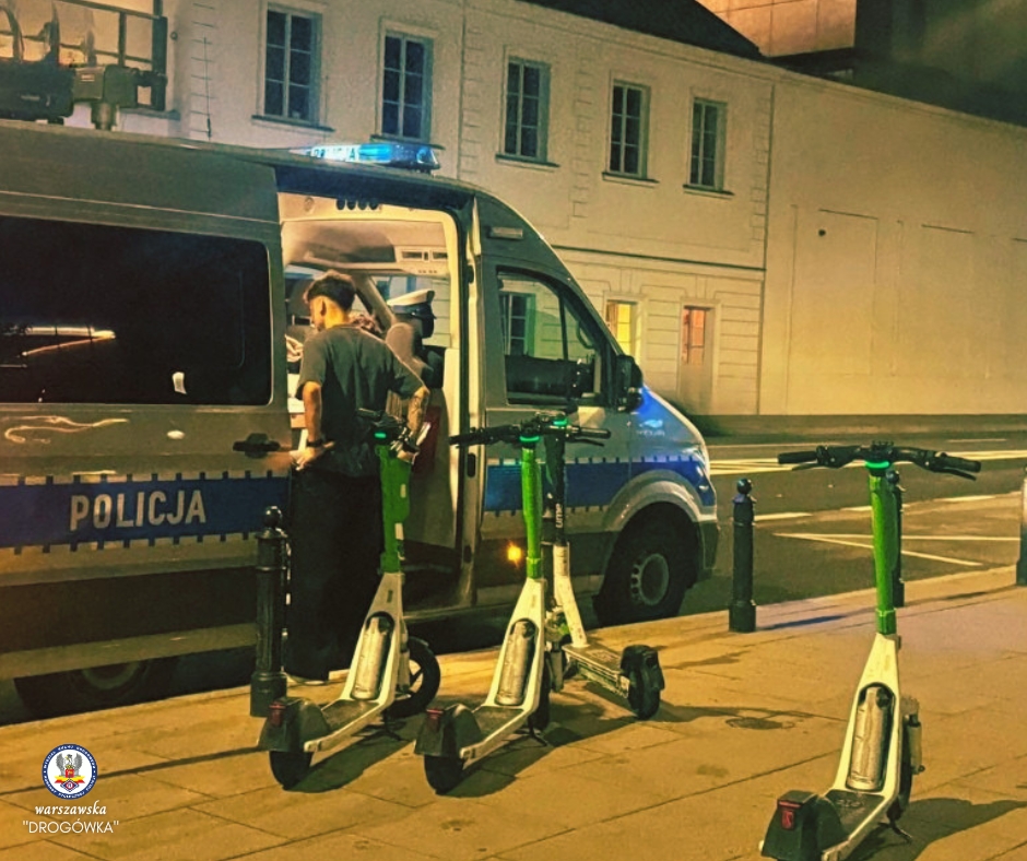 Mężczyzna w ciemnej koszulce przegląda dokumenty w otwartej furgonetce policyjnej, obok stoją elektryczne hulajnogi.