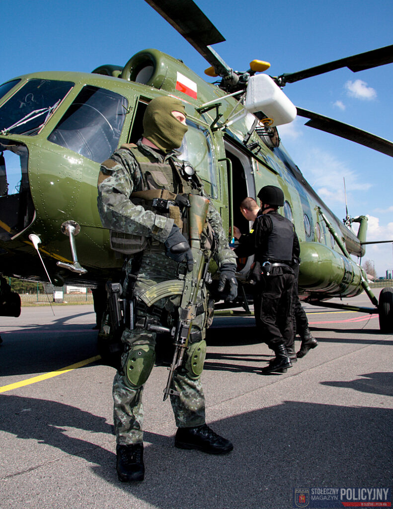 Uzbrojony policjant w kamizelce ochronnej stoi obok śmigłowca wojskowego, podczas gdy inni funkcjonariusze wchodzą do maszyny.