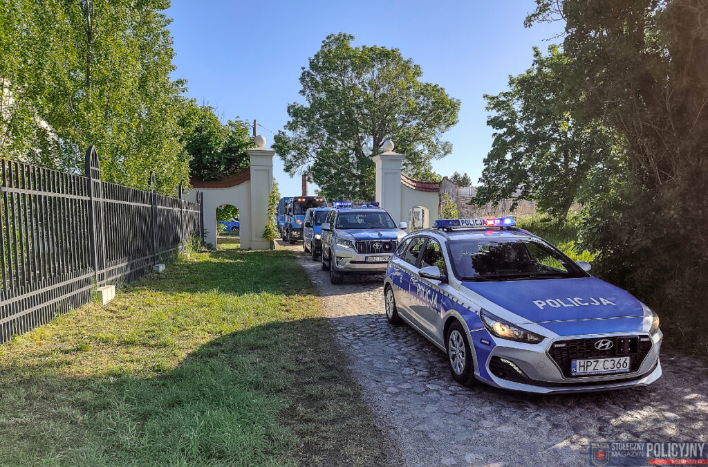 Policyjny radiowóz z włączonymi sygnałami świetlnymi wjeżdża przez bramę otoczoną zielenią.