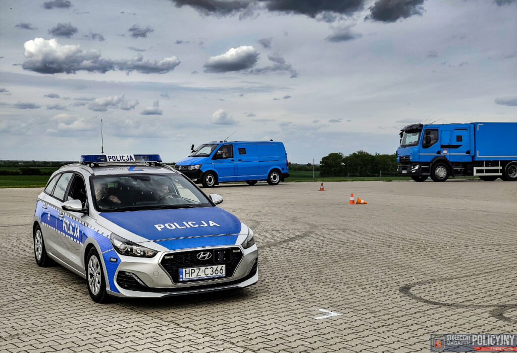 Policyjny samochód osobowy z niebieskimi oznaczeniami stoi na placu, obok dwóch niebieskich pojazdów dostawczych.