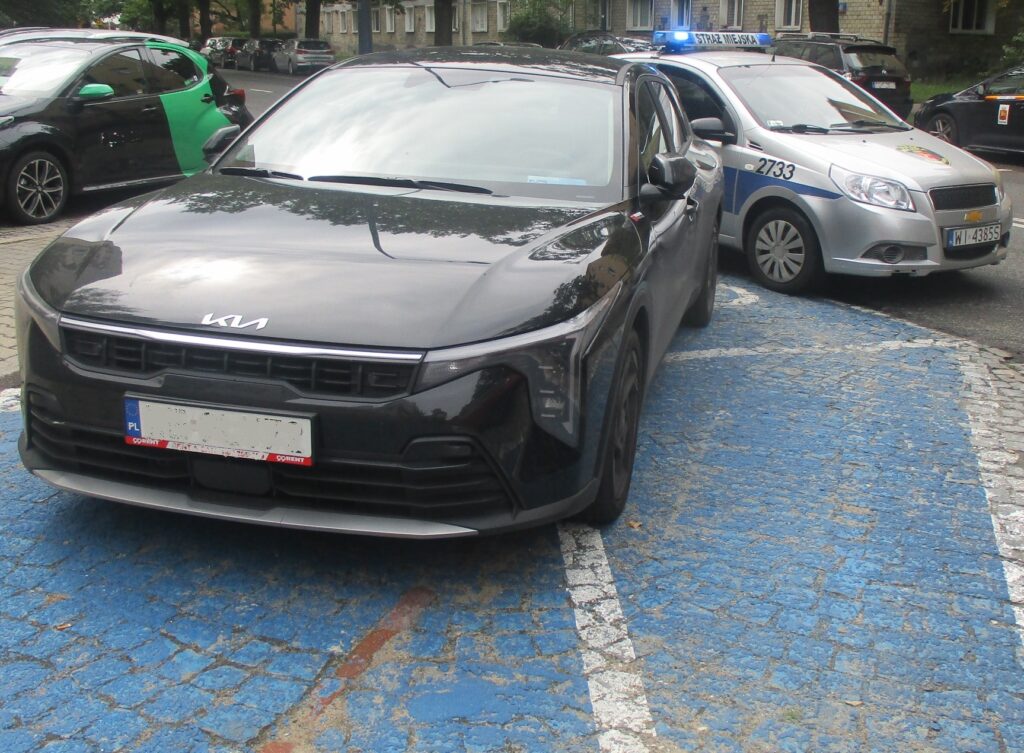 Czarny samochód osobowy zaparkowany na niebieskim miejscu parkingowym, obok radiowóz policyjny z włączonymi sygnałami świetlnymi.