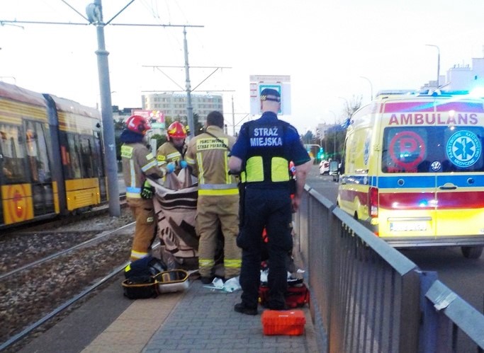 Grupa strażaków oraz strażników miejskich udziela pomocy mężczyźnie na przystanku tramwajowym. W tle widoczna karetka.