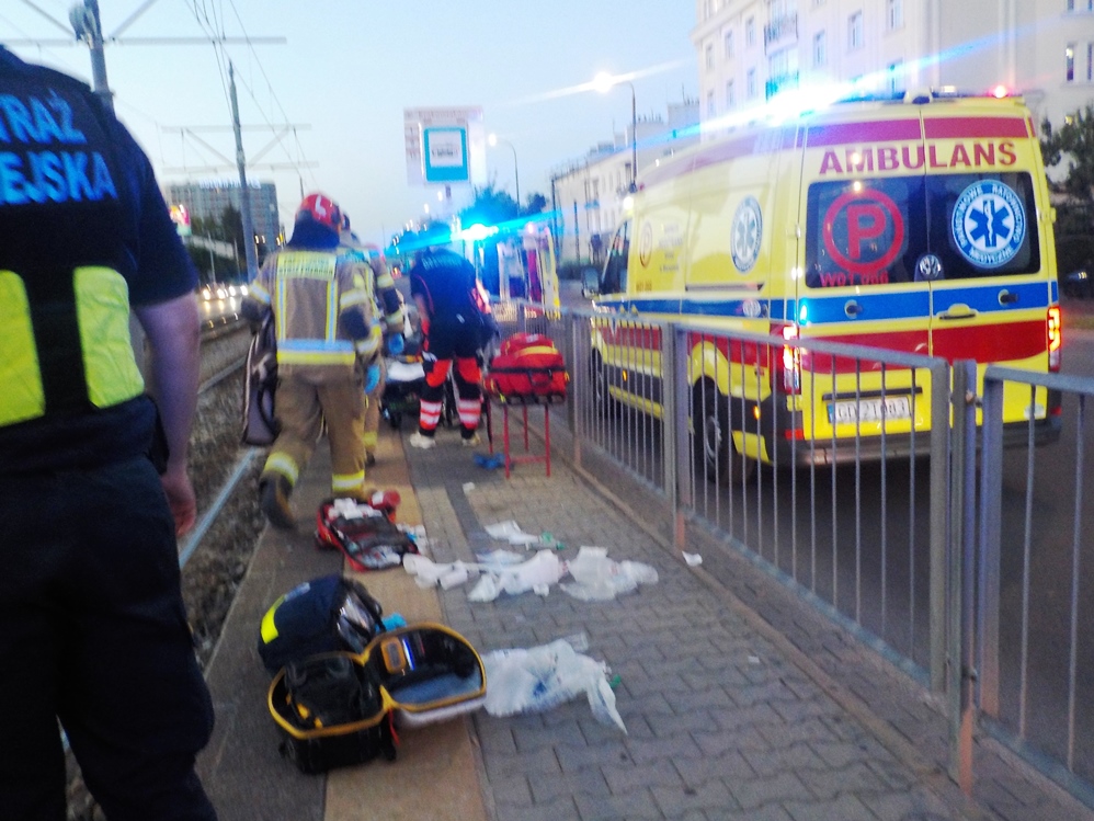 Ratownicy medyczni udzielają pomocy mężczyźnie leżącemu na przystanku tramwajowym, w tle widać karetki.