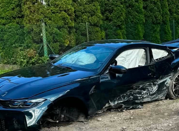 Czarny samochód osobowy z uszkodzeniami leży na poboczu drogi, otoczony zielonymi drzewami i siatką.