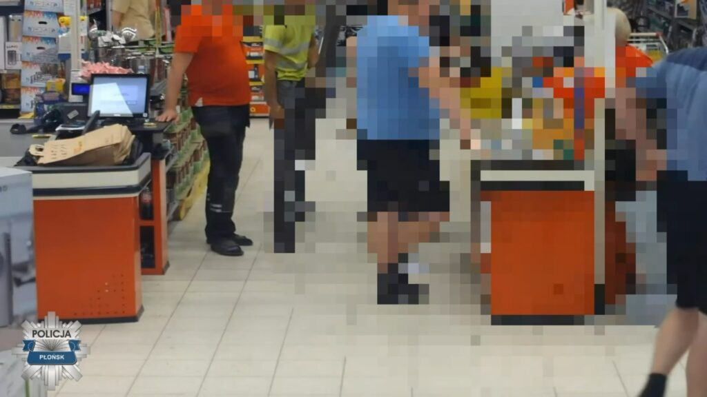 Mężczyzna w niebieskiej koszulce stoi przy ladzie w sklepie, obok niego dwóch innych mężczyzn, w tle regały z towarem.