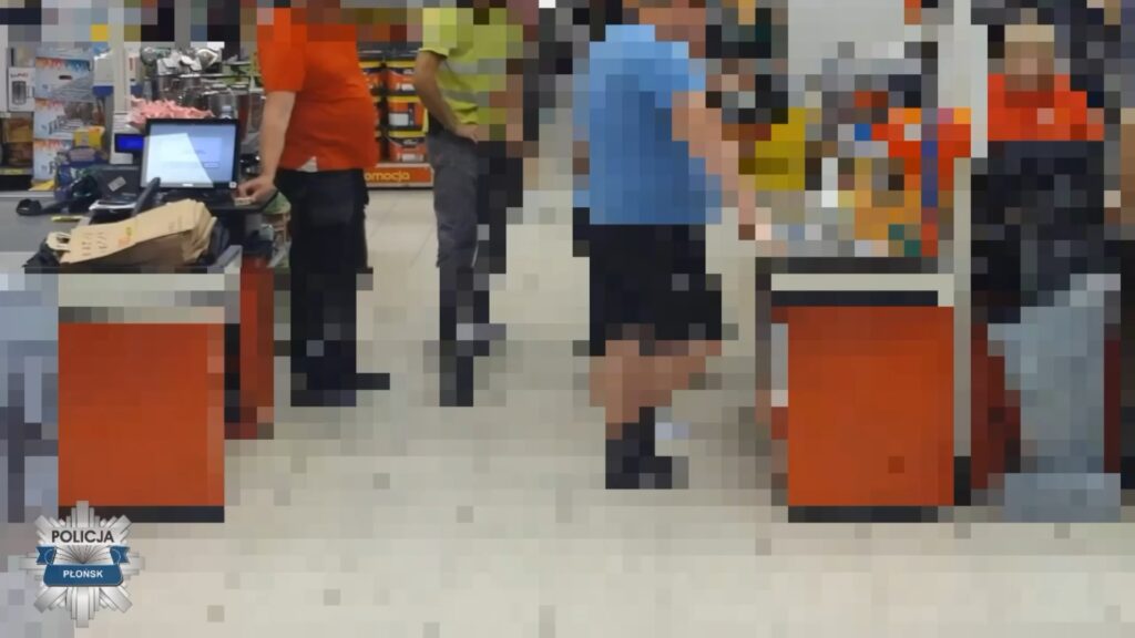 Mężczyzna w niebieskiej koszulce stoi przy kasie w sklepie, obok niego dwie osoby w odzieży roboczej.