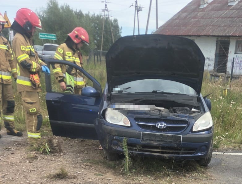 Grupa strażaków w mundurach sprawdzająca niebieski samochód osobowy z otwartą maską na poboczu drogi.