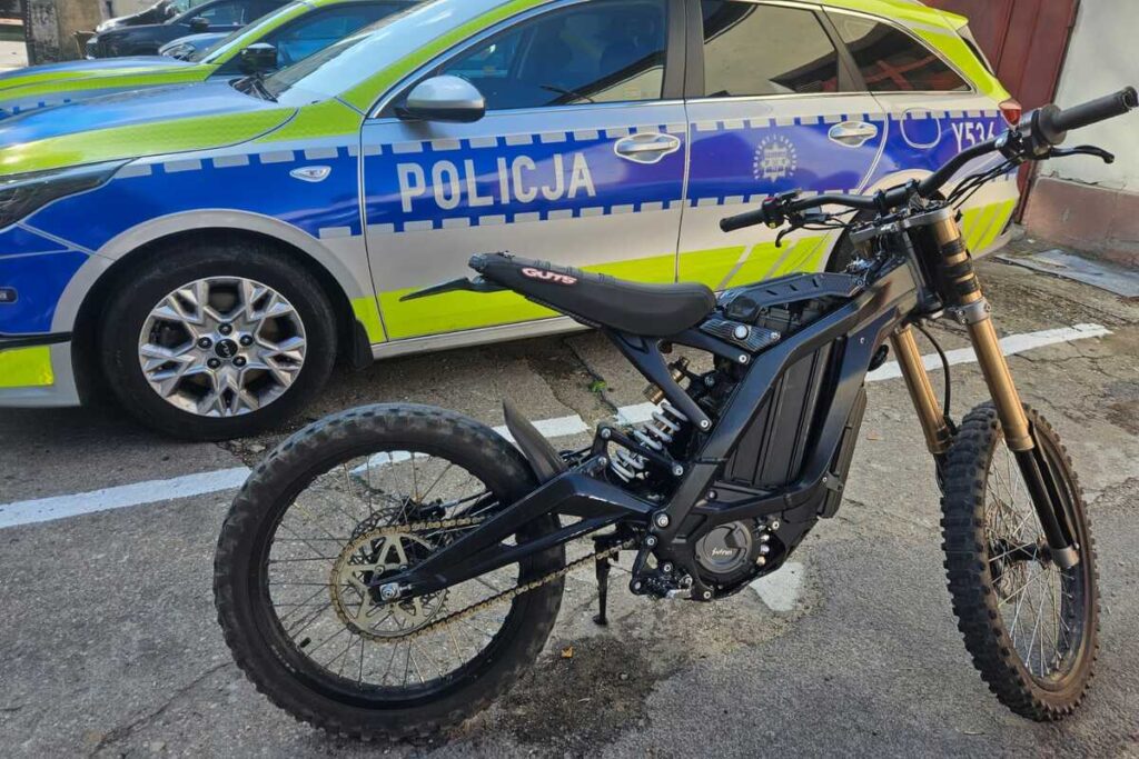 Czarny rower elektryczny stoi obok policyjnego samochodu z niebiesko-zielonym oznakowaniem na parkingu.