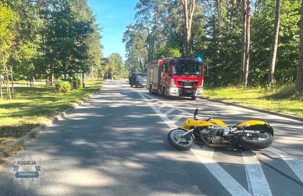Na drodze leży przewrócony żółty motocykl, obok przejeżdża czerwona ciężarówka strażacka w otoczeniu drzew.