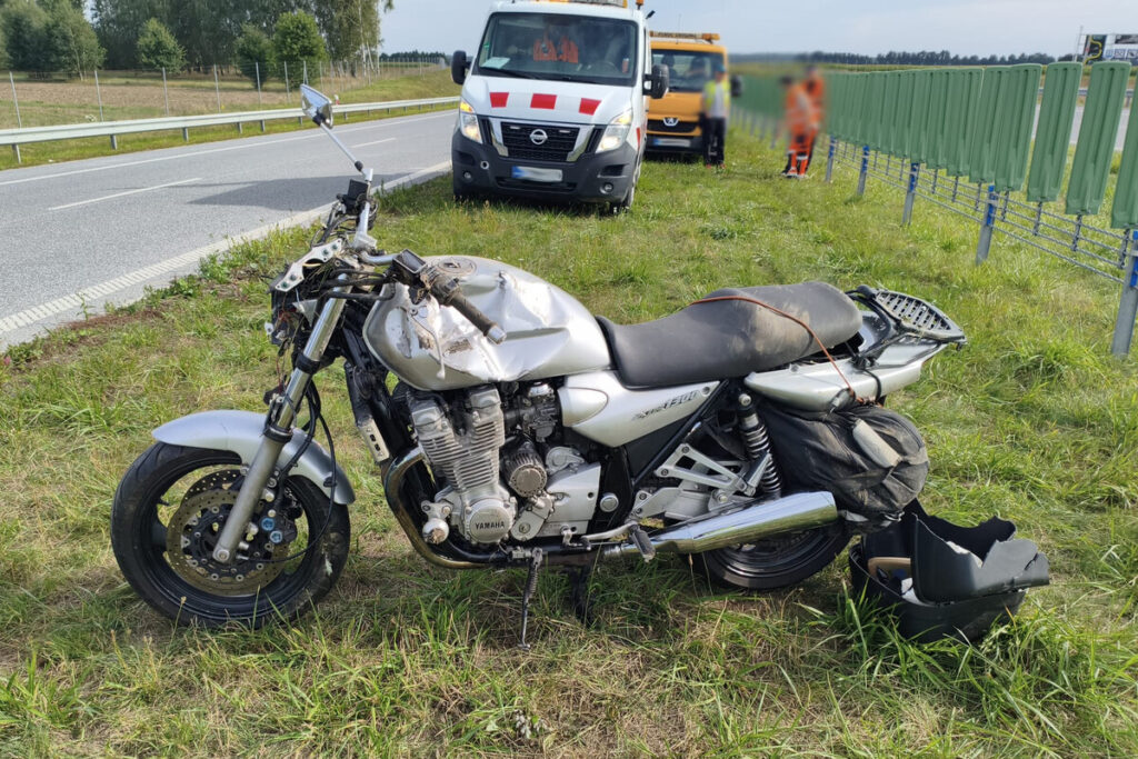 Zniszczony motocykl leży na trawie obok drogi, a w tle widać samochody służb ratunkowych.