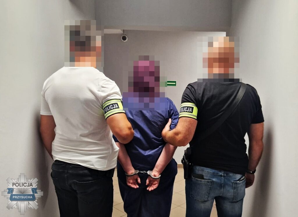 Kobieta w kajdankach prowadzona przez dwóch policjantów w korytarzu budynku.