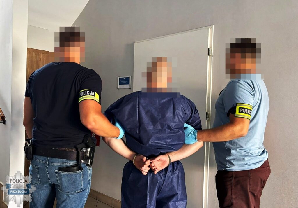 Mężczyzna w niebieskim stroju medycznym jest prowadzony przez dwóch policjantów w mundurach, w korytarzu budynku.