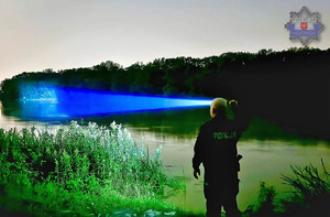 Policjant z latarką o niebieskim świetle przeszukuje brzeg rzeki w nocy, otoczony roślinnością.