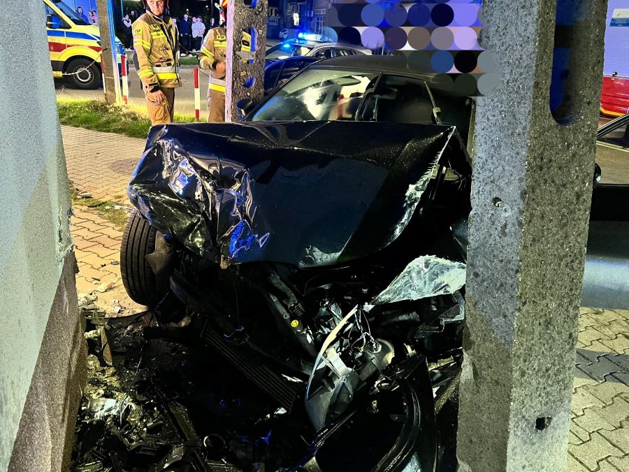 Czarny samochód osobowy wjechał w ścianę budynku, zniszczona przednia część pojazdu, w tle interwencja służb ratunkowych.