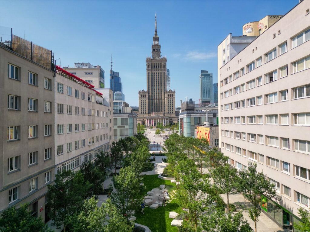 Nowoczesne budynki otaczają zielony skwer z ławkami, w tle widać Pałac Kultury i Nauki.