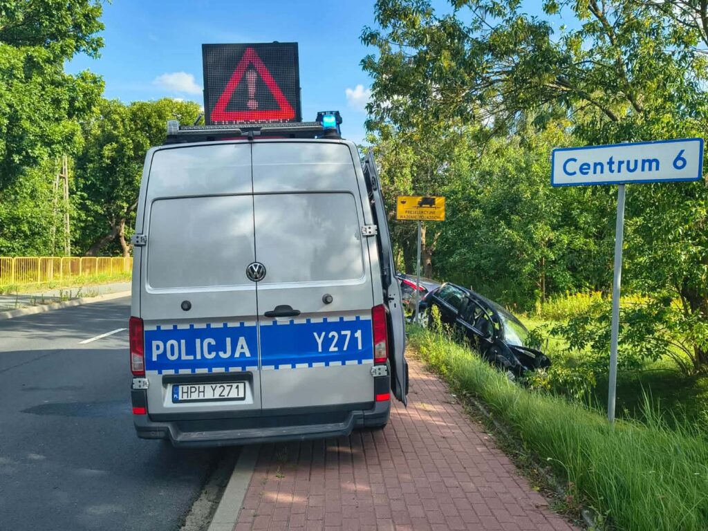 Policyjny van z włączonym sygnalizatorem stoi przy drodze, obok leżącego w rowie uszkodzonego samochodu.