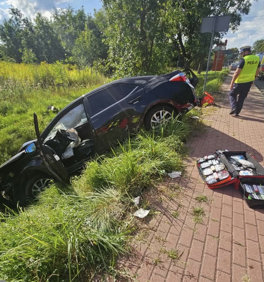 Czarny samochód osobowy w rowie, obok stoi policjant w kamizelce odblaskowej, a na ziemi leży torba z wyposażeniem.