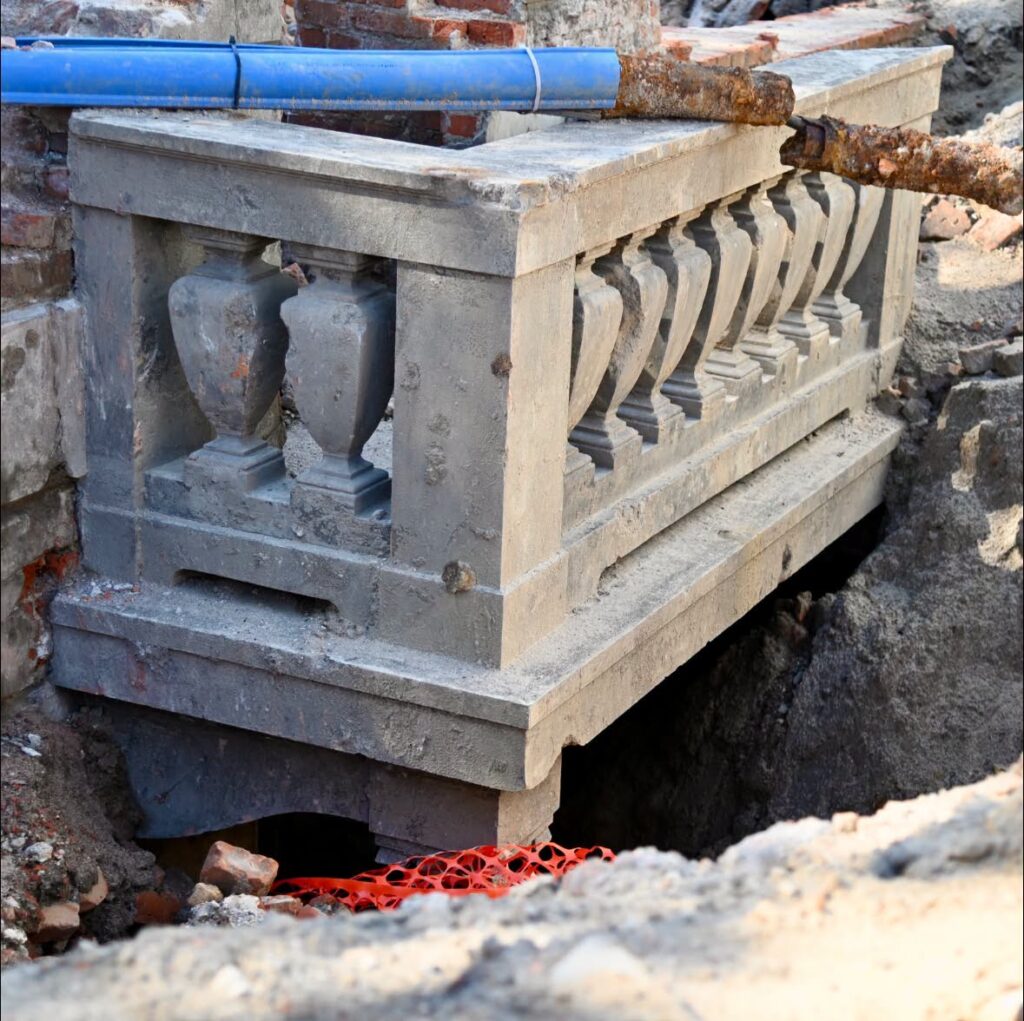 Odsłonięta betonowa balustrada z dekoracyjnymi urnami, otoczona wykopem budowlanym.