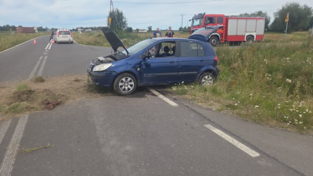 Niebieski samochód osobowy z uniesioną maską stoi na poboczu drogi, obok zespół ratunkowy oraz inne pojazdy.