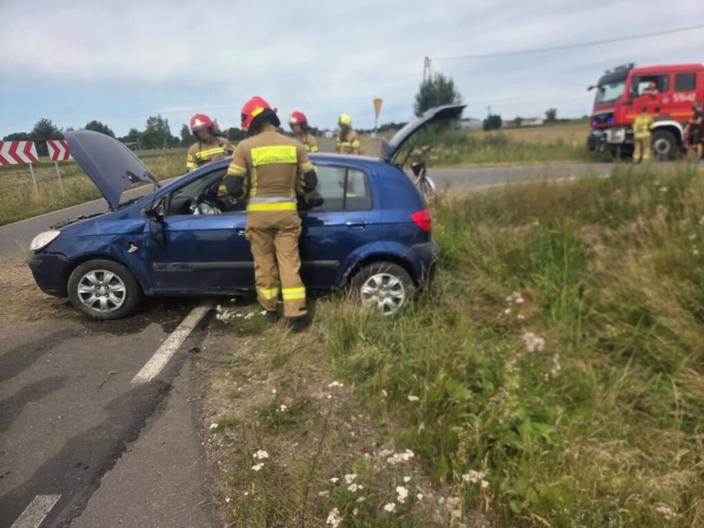 Strażacy z interwencyjnym sprzętem pomagają przy uszkodzonym niebieskim samochodzie na poboczu drogi.