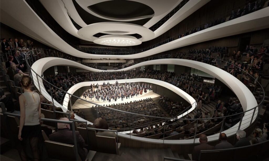 Sala koncertowa z widownią wypełnioną ludźmi, orkiestra na scenie, nowoczesna architektura z falistymi elementami.