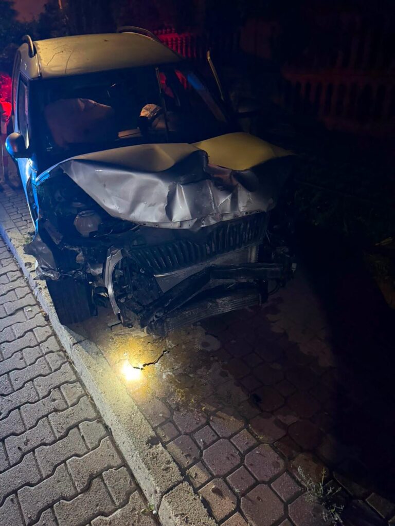 Uszkodzony samochód osobowy z wgniecionym przodem stoi na chodniku oświetlonym przez latarnię, w nocy.