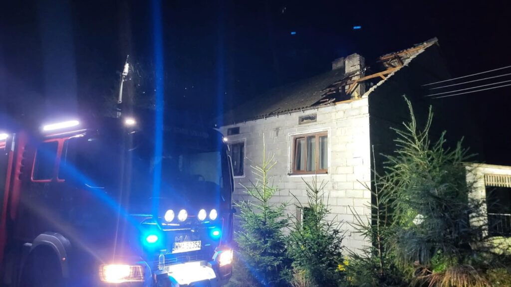 Czerwony wóz strażacki z włączonymi sygnalizatorami przed uszkodzonym budynkiem z zerwaną dachówką w nocnym otoczeniu.