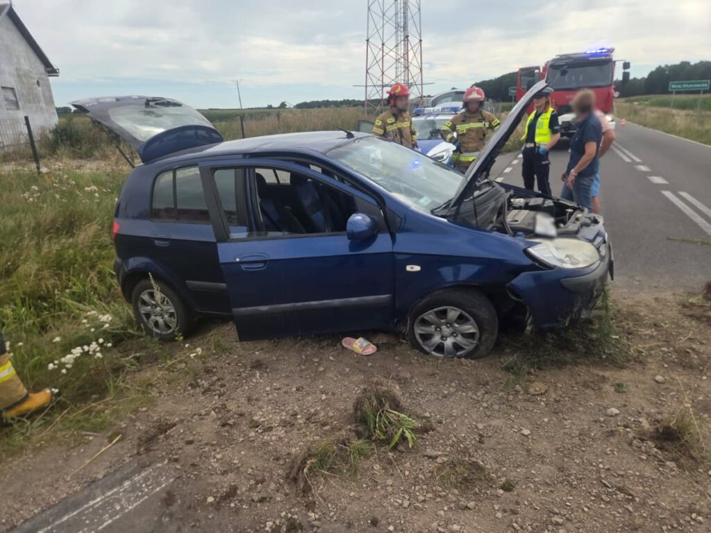 Niebieski samochód osobowy stoi na poboczu drogi, a obok grupka ratowników w mundurach omawia sytuację.