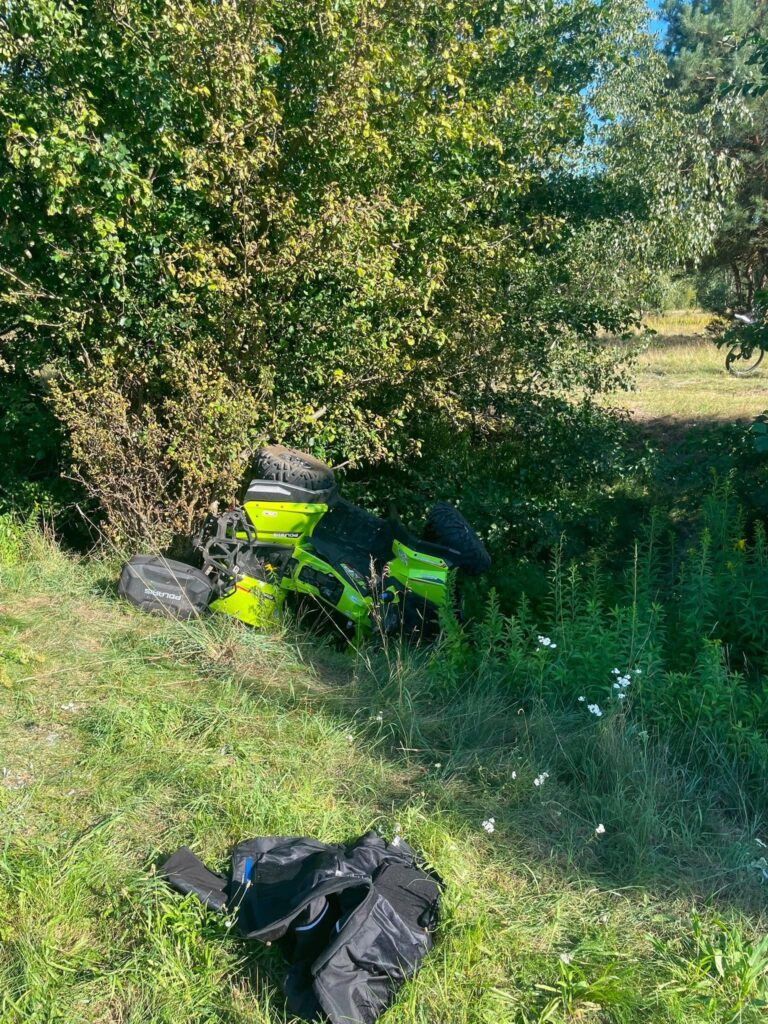 Zielony quad przewrócony na bok w zaroślach, obok leży czarna kurtka motocyklowa na trawie.