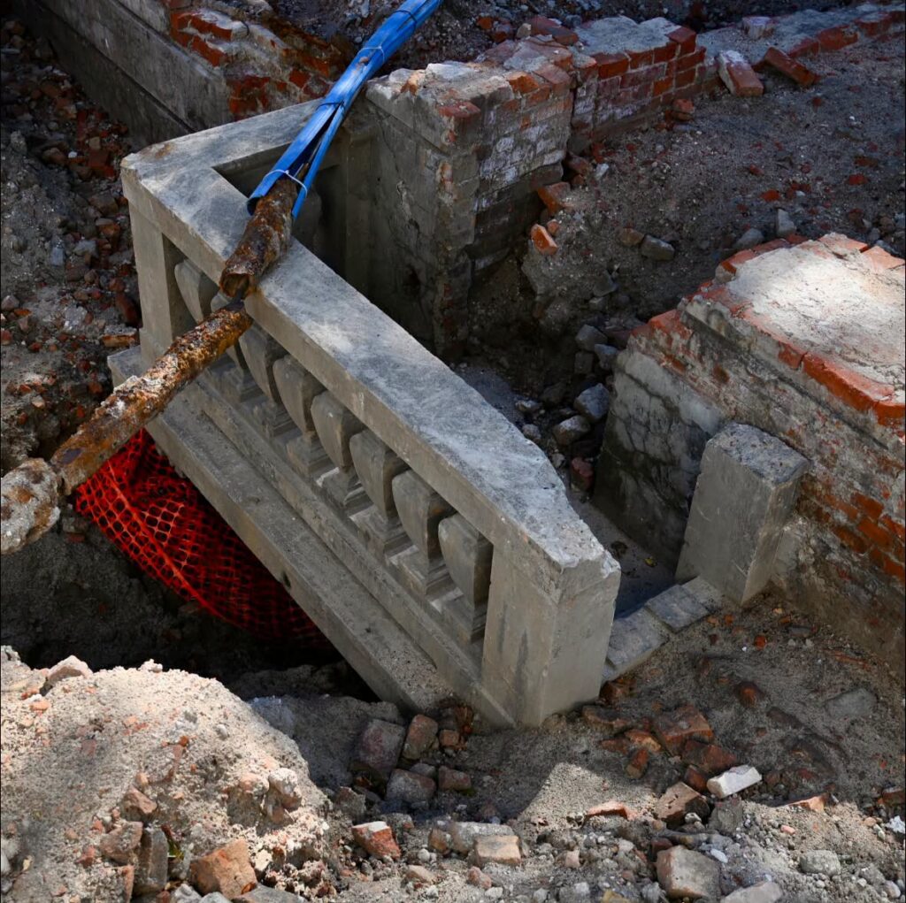 Fragment betonowej balustrady odkryty podczas remontu, otoczony cegłami i ziemią w wykopie budowlanym.