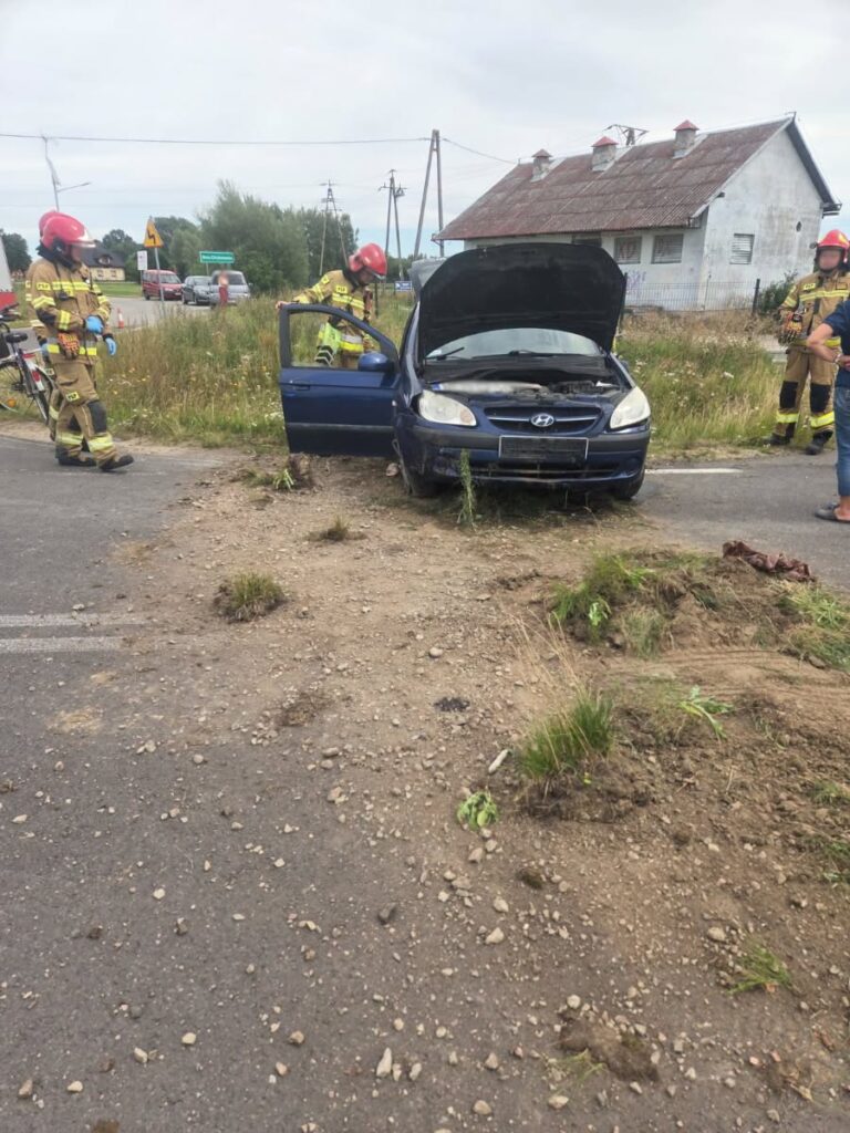 Strażacy w kaskach badają niebieski samochód osobowy z otwartą maską na poboczu drogi, obok starego budynku.