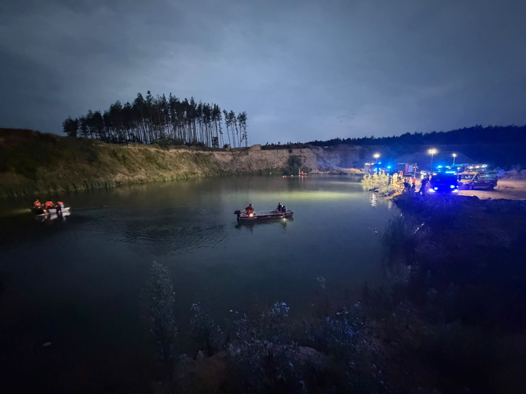 Grupa ratowników na łodziach przeszukuje wodę w kamieniołomie w nocy, otoczona pojazdami z włączonymi sygnałami świetlnymi.