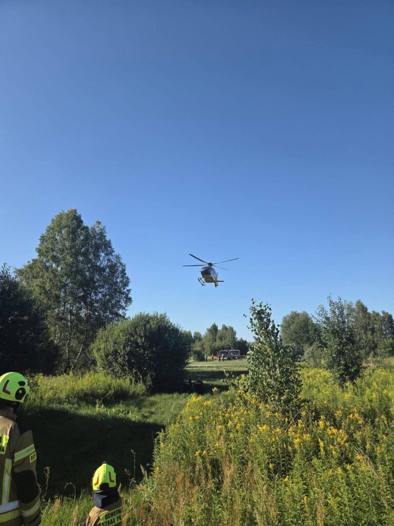 Śmigłowiec ratunkowy przelatujący nad łąką, obok strażacy w hełmach obserwujący akcję ratunkową.